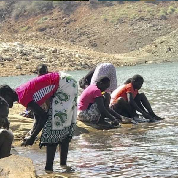 Women and children crouched along a rocky riverbank, washing clothes where the water meets the stones.