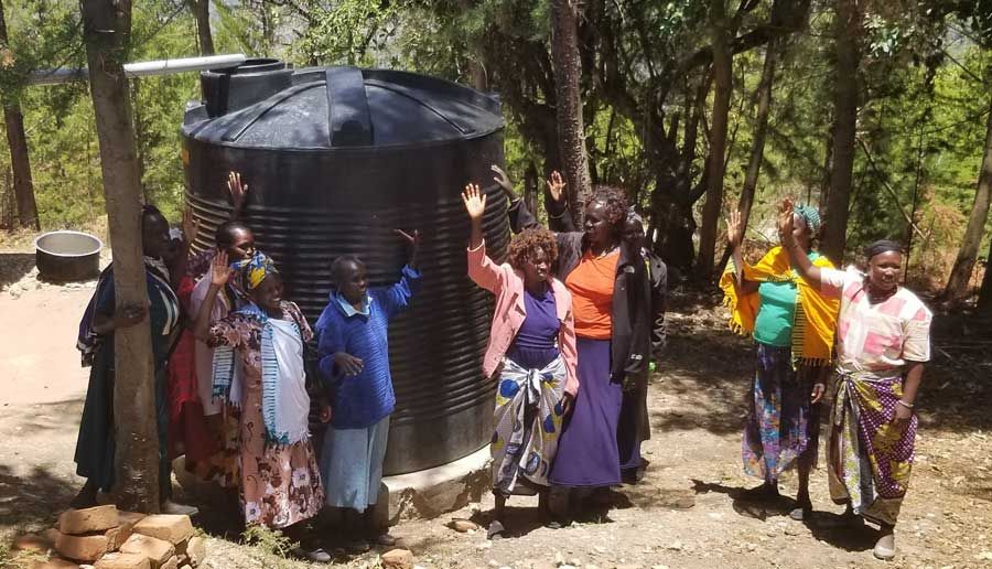 Group of women standing around a large black water tank with their hands raised.