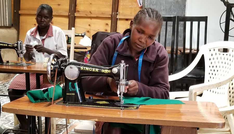Young woman guiding green fabric through a treadle sewing machine, with a second student working behind her.